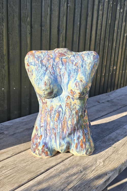 Keramisk skulptur af en kvindelig torso med abstrakt, flerfarvet glasur stående på en træbænk foran en sort væg. (1)
