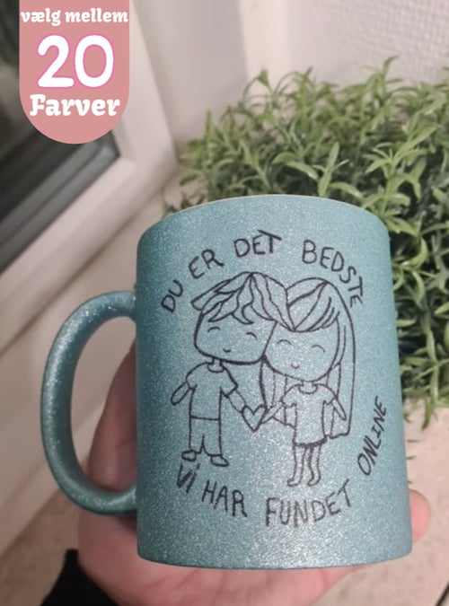En hånd holder en glitrende turkis kaffekop med en tegning af en dreng og pige, der holder hænder. Teksten på koppen lyder: "Du er det bedste vi har fundet online." Tekst i øverste venstre hjørne: "vælg mellem 20 farver." (1)