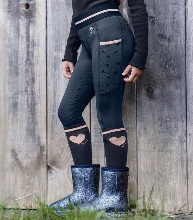 Barn iført sorte ridebukser med pink detaljer, sorte strømper med lyserøde hjerter og glitrende støvler stående foran et træhegn.