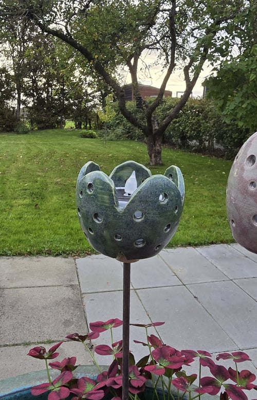 Unik grøn keramisk havefakkel med huller på en metalstang, placeret blandt planter og en græsplæne med træer i baggrunden. (1)