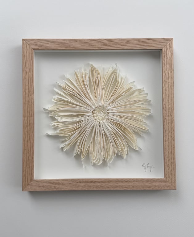 Billede af en vægdekoration med et fyrretræsramme, der indeholder en skulptur af en blomst lavet af foldede hvide papirsider.