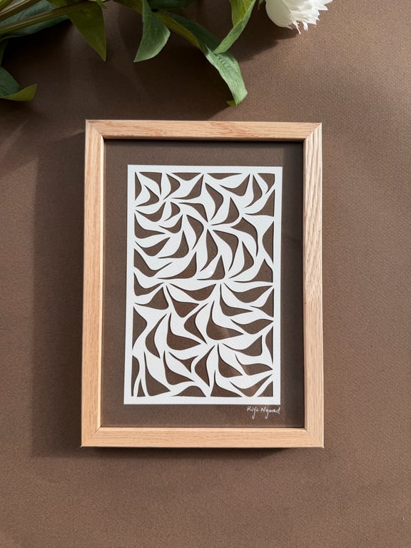Indrammet papercut-kunst med abstrakte bladformer i hvidt på brun baggrund, placeret på brunt underlag med grønne blade og en hvid blomst delvist synlig øverst.