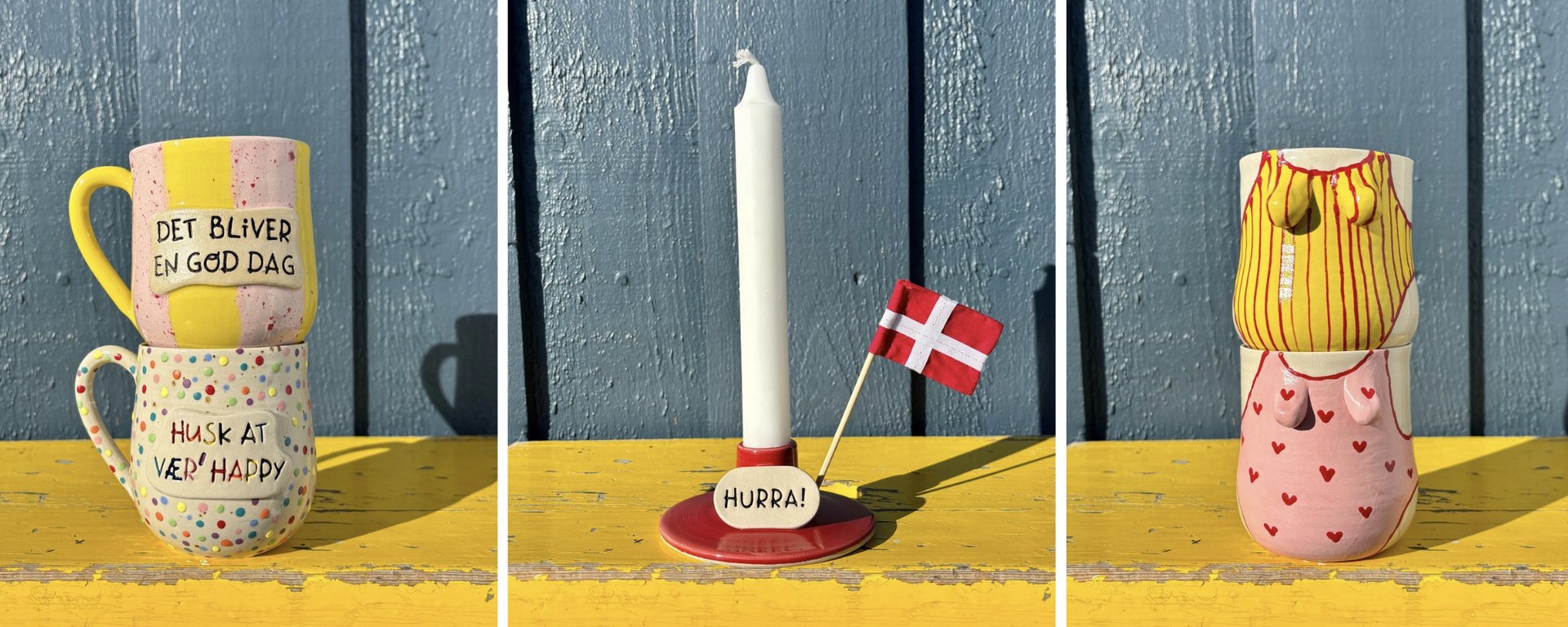 Stack af tre hvide keramiske skåle med teksten "Slikskålen", to hvide keramiske krus med teksten "Smil smitter" og et hvidt keramisk fad med teksten "Hurra" og danske flag, alle med farvede prikker, placeret på en gul hylde foran en blå væg.