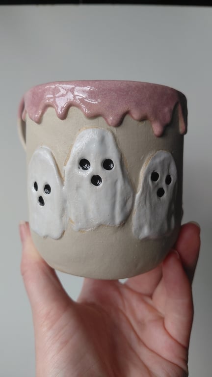 Hånd holder en keramikkop med pink glasur på kanten og tre hvide spøgelsesfigurer med sorte øjne og mund som dekoration.