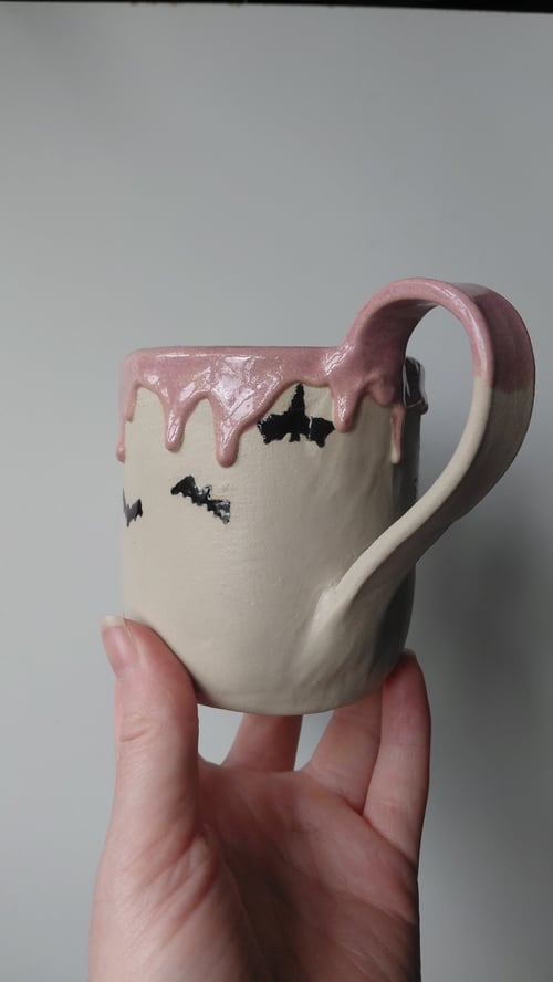 En hånd holder en keramisk kop med en lysegrå bund og pink glasur, der drypper ned fra toppen. Koppen har sorte flagermus-motiver rundt om siderne. (3)