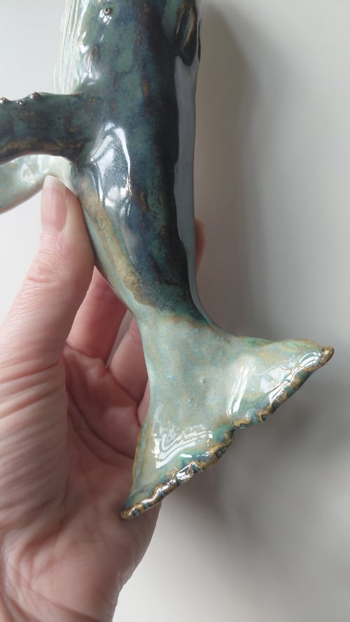 Hånd, der holder en keramisk fisk med glaseret blågrønne farver, fokus på fiskens hale. (4)