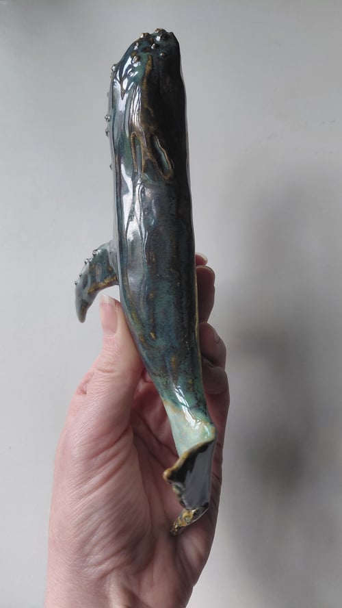 Hånd, der holder en blågrøn glasurkeramikfigur formet som en hval med finner. (8)