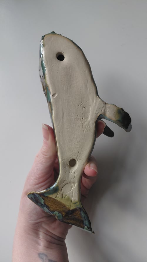 Hånd holder en keramisk hvalfigur set fra undersiden med to huller og synlige glasurdetaljer. (9)