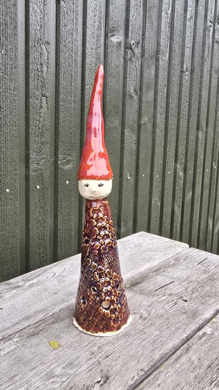 Keramisk nissedekoration med rød spids hat, rund hvidt ansigt og tekstureret brun krop, placeret på et træbord foran en grøn trævæg.