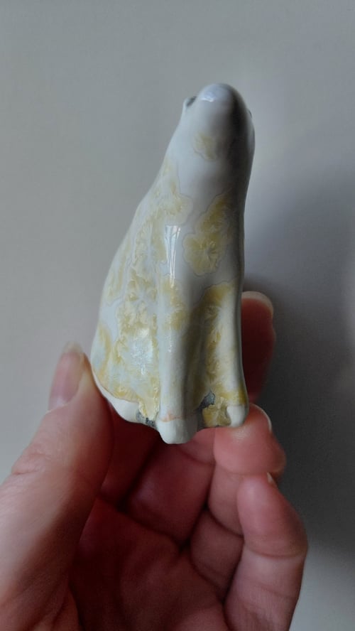 Hånd, der holder en lille, hvid og gul, glaseret porcelænsfigur formet som en siddende hund. (2)