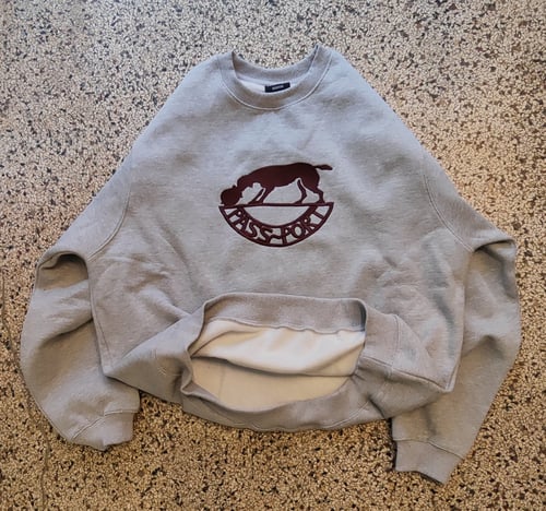 Grå sweatshirt med rund hals og logo af en hund over teksten "PASSPORT" på fronten, lagt fladt på en stenmønstret gulv. (2)