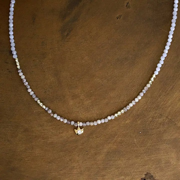 Halskæde med små perler i lyse lilla nuancer og gulddetaljer samt en lille guldfugl med en perle ved midten, liggende på en træflade.