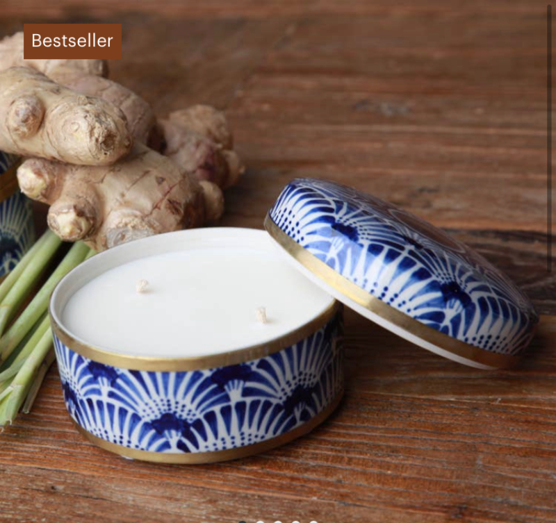 Dekorativt hvidt duftlys med to væger i blå-hvidt mønstret metalæske med låg, placeret på træbord med frisk ingefær og citrongræs ved siden af. (2)