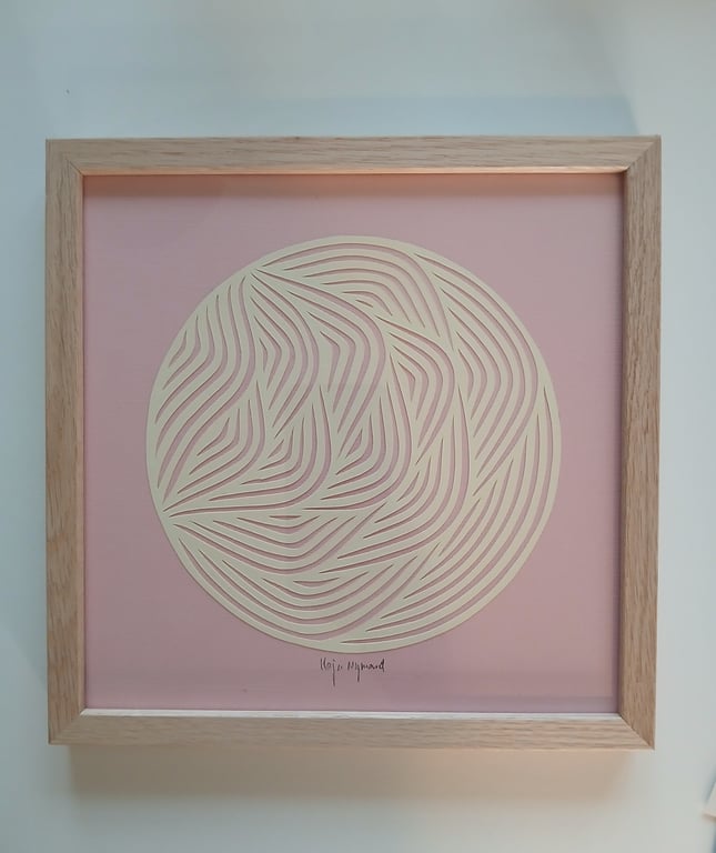 Abstrakt papirkunst i en lys treramme, med bølgende, udskårne mønstre i en cirkel på en lyserød baggrund.