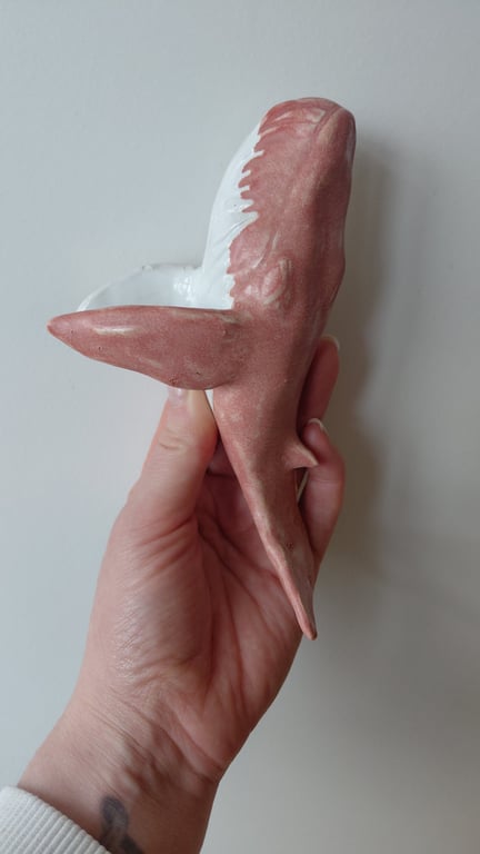 Hånd, der holder en lille keramisk figur formet som en hvalhaj, malet i rødt og hvidt.