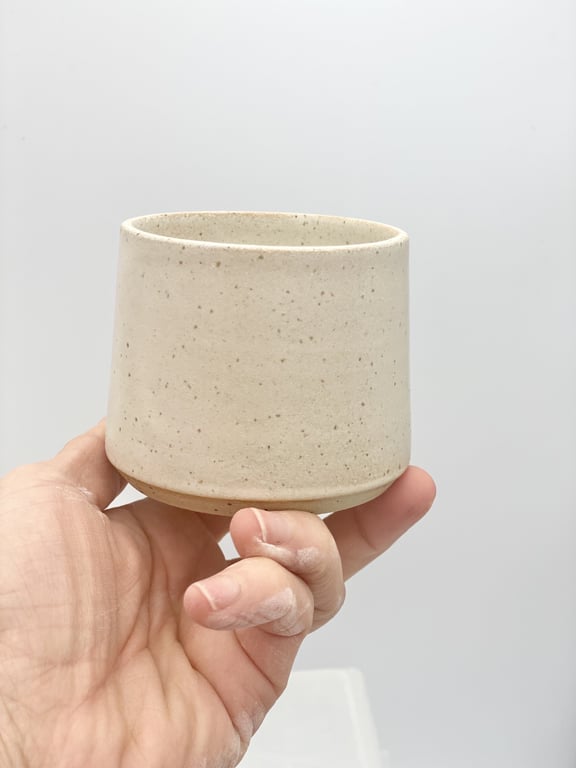 Hånd der holder en beige, keramik kop med enkle, små prikker mod en lysegrå baggrund.