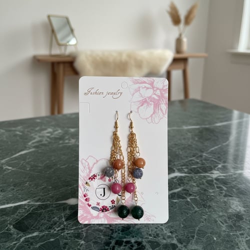 Par af hængende modeøreringe med flerfarvede perler i pink, blå, brun og grøn, hængende på guldfarvede kæder, vist på hvidt kort med blomsterdekoration, placeret på grøn marmorbord. (1)