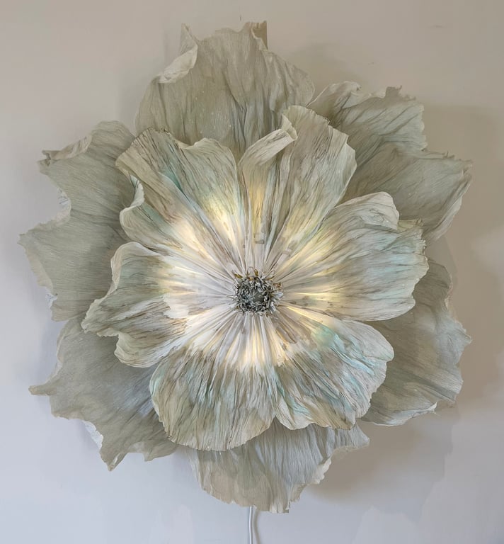 Stor, kunstnerisk vægdekoration formet som en åben blomst med flere lag af bølgede, lyse blade og blødt lys, der skinner igennem midten.