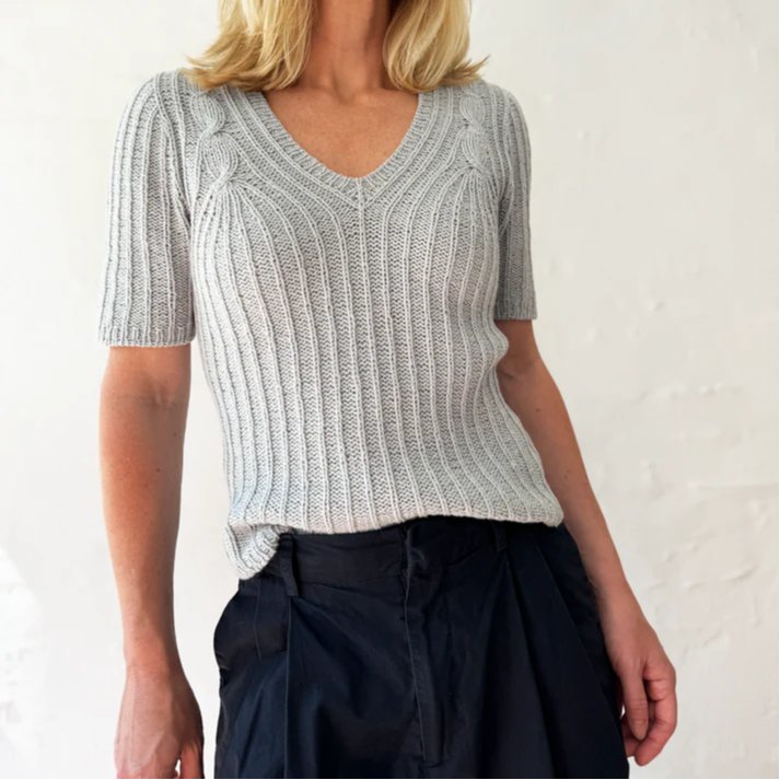 Kvinde iført strikket lysegrå kortærmet trøje med V-hals og sort nederdel eller bukser.