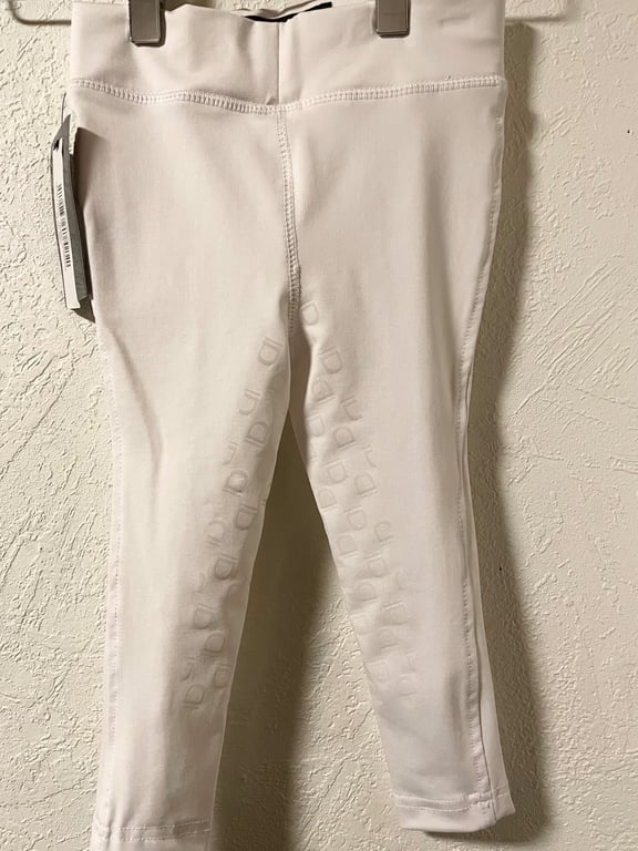 Billede af et par hvide ridebukser til børn med antislip på indersiden af lårene, hængende på en klemme mod en hvid væg.