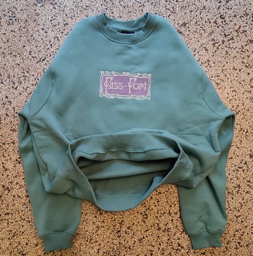 Grøn sweatshirt med rund hals og lilla mærke med teksten "Pass-Port" placeret på forsiden, lagt fladt på en marmoreret gulvflade. (2)