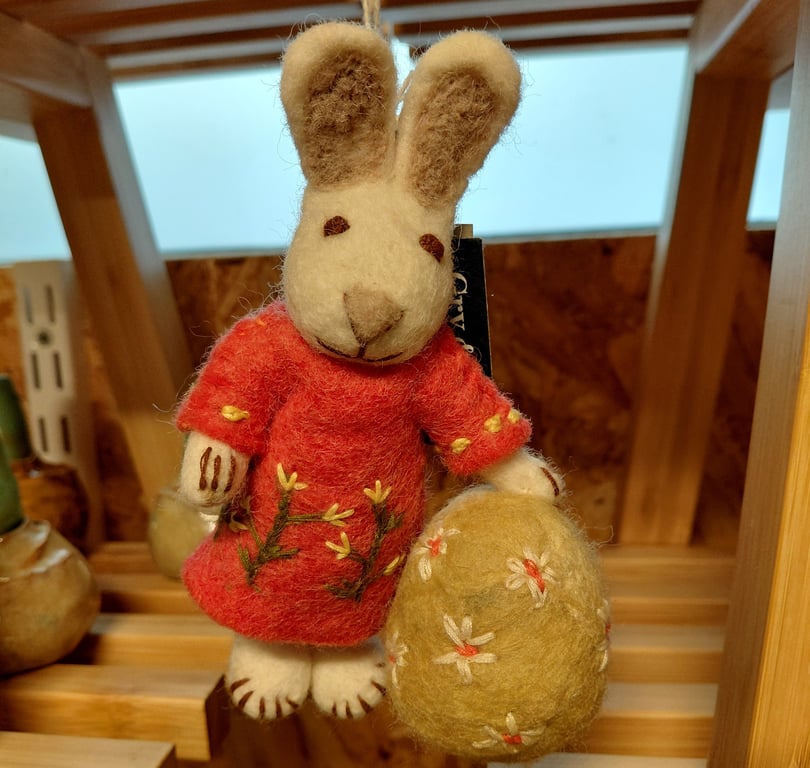 Filtet påskehare iført rød kjole med broderede blomster, der holder et stort gult æg med hvide og orange blomsterdekorationer.