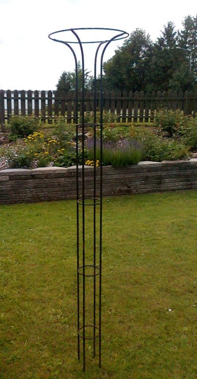 Sort metal plantehumørk med en rund åbning øverst, placeret på græsplæne i en have med blomsterbed og træhegn i baggrunden.