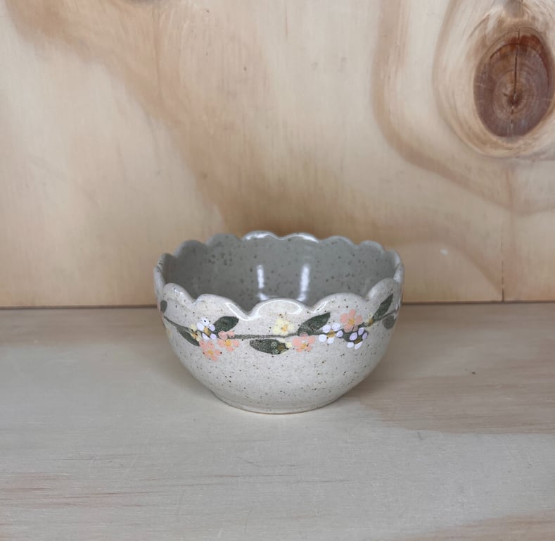 Keramisk skål med bølget kant og håndmalet blomsterdekoration, placeret på lyst træbord mod lys trævæg.