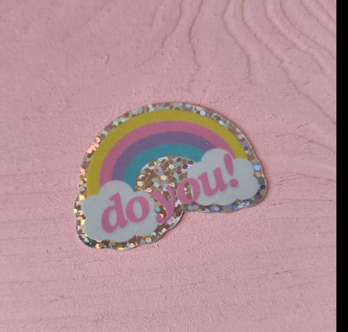 Sticker med glitrende kant, der forestiller en regnbue med farverne gul, lyserød, lilla og blå samt to hvide skyer, hvor der står "do you!" i pink tekst, på en lyserød overflade. (2)