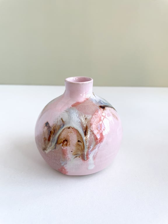 Lille rund keramikvase i lyserød med abstrakte brune, hvide og blå glasurdetaljer, placeret på en hvid overflade.