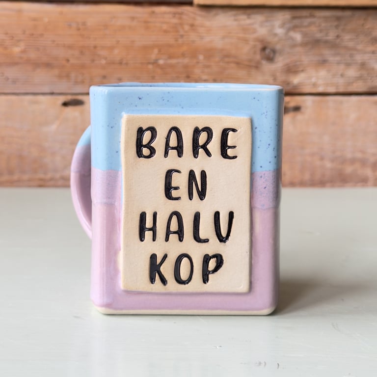 Kop med pastelblå og lyserød farve med teksten "Bare en halv kop" foran en træbaggrund.