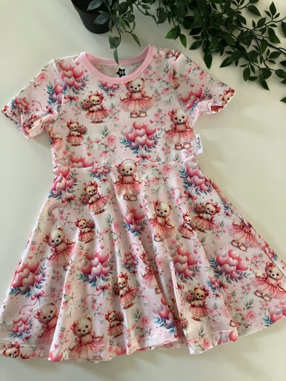 Lyserød kjole til børn med mønster af bjørne i kjoler, balloner og blomster, lagt ud på hvid overflade med grønne planter i baggrunden.