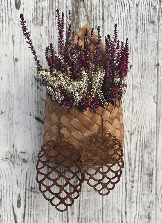 Flettet kurv med lilla og hvide lyngblomster hængende på hvidt, slidt trægulv med rustne metalkogledekorationer foran.