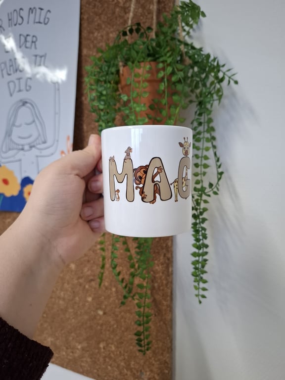 Hånd, der holder en hvid kaffekop med bogstaver dekoreret som dyr foran en grøn, hængende plante og en opslagstavle med en tegning og dansk tekst.
