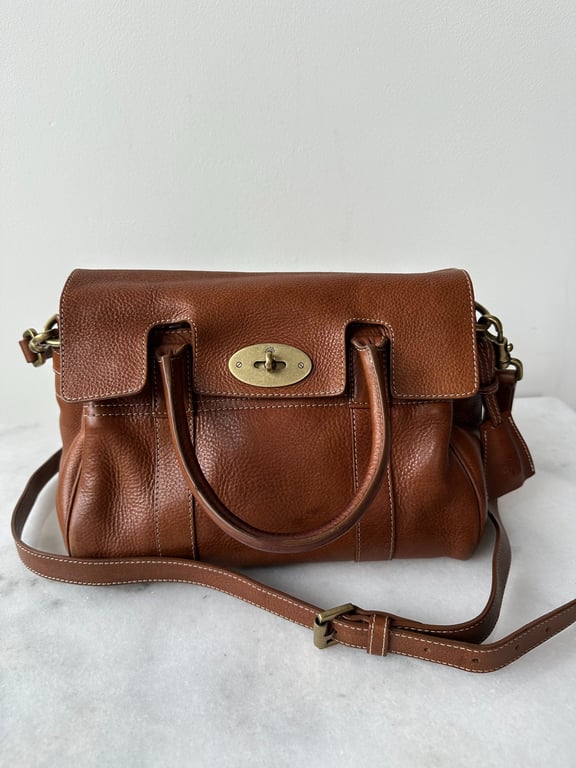 Brun lædertaske med håndtag, justerbar skulderrem og guldfarvet lås på hvid baggrund.