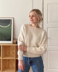 Kvinde i beige striktrøje og blå jeans står i lyst rum med træreol og kunstværk i baggrunden. (1)