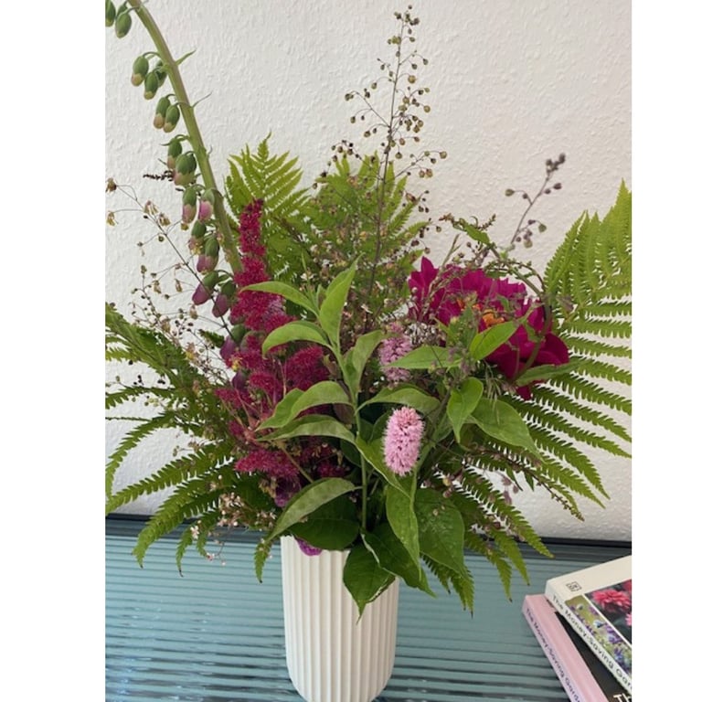 Buket med grønt løv og blomster i forskellige lilla og pink nuancer i en hvid, ribbet vase på et blåt bord ved en hvid væg.
