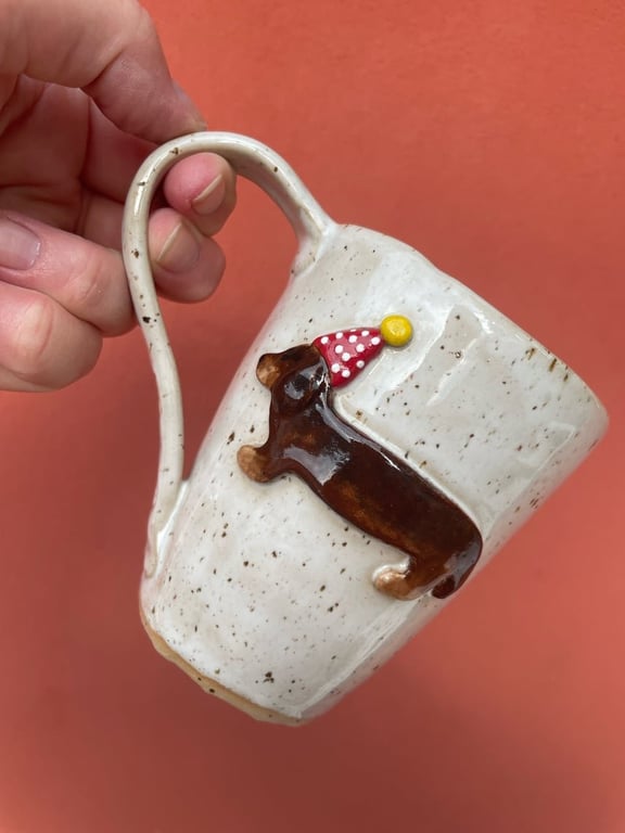 Hånd der holder hvidt keramikkrus med en dekoreret gravhund med rød nissehue på mod orange baggrund.