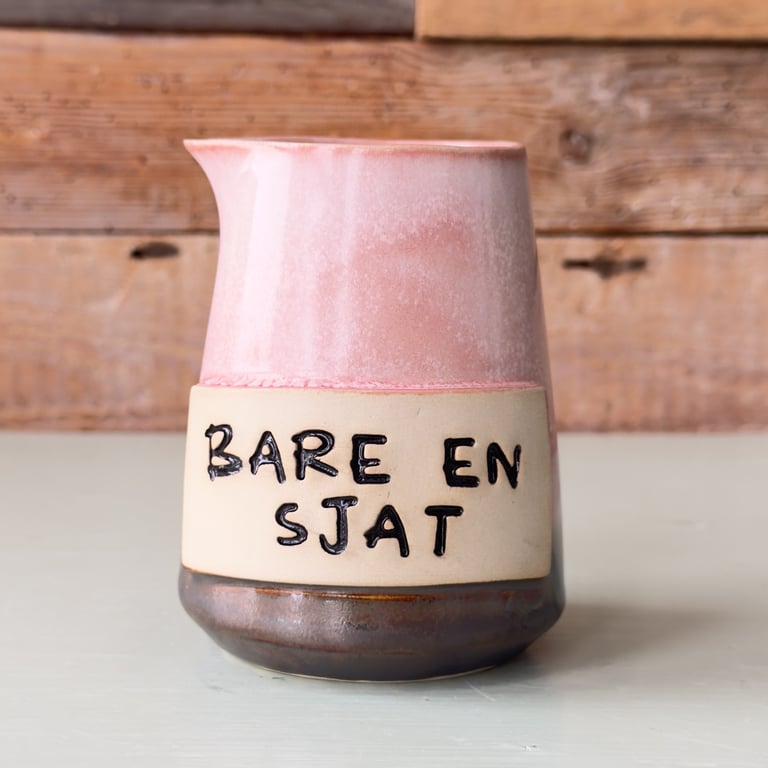 Keramisk kande med tre farver: lyserød, beige og brun, med teksten "BARE EN SJAT" skrevet i sort på beige del. Kanden står på en lys overflade foran en trævæg.