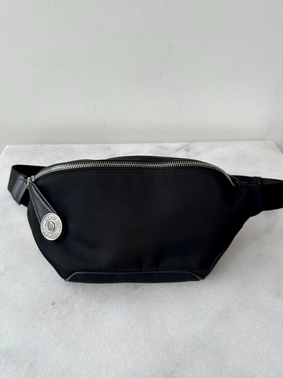 Sort bæltetaske med sølvfarvet lynlås og rund metalmærke på hvid marmorflade.