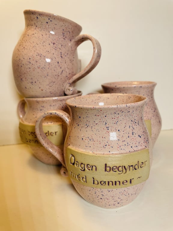 Billede af fire keramikkrus med lyserød og blå stænk, hvoraf to har en gul mærkat med teksten "Dagen begynder med bønner".