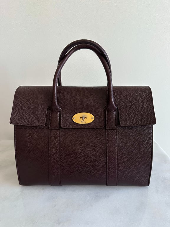 Brun skindtaske med dobbelt håndtag og gyldent spænde foran, placeret på en hvid overflade mod en hvid væg.