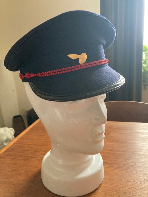 Blå kasket med rødt bånd og gul emblem, placeret på en hvid mannequin-hoved, på et træbord indendørs. (2)