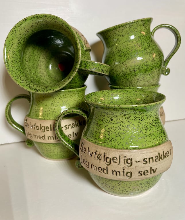 Grøn, keramisk kop med hank og dansk tekst "Selvfølgelig - snakker jeg med mig selv" på beige bånd. Flere kopper stablet op mod hvid baggrund.
