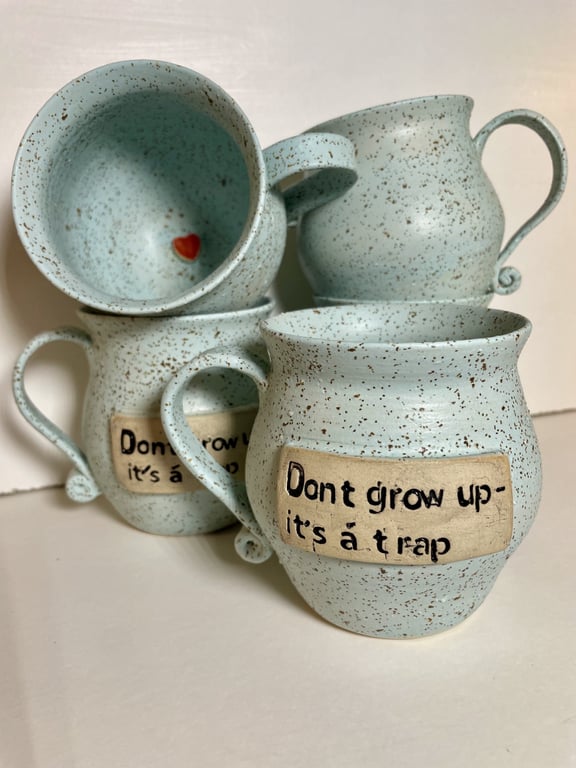 Fire lyseblå keramikkrus med brune pletter, hvoraf to har teksten "Don't grow up - it's a trap" og et krus viser en lille rød hjertefigur indeni.