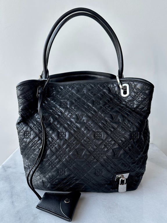 Sort quiltet læderhåndtaske med håndtag, sølvfarvet lås og lille tilbehørspung, stående på hvid overflade.