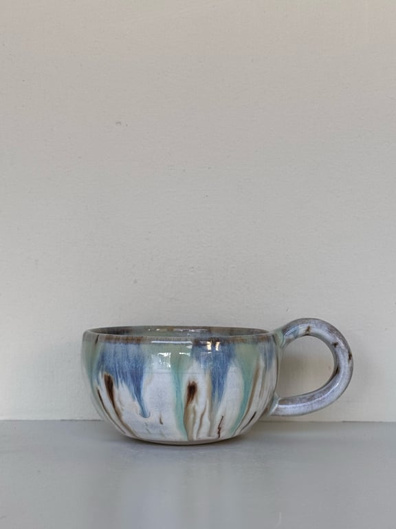 Keramikkop med blå, grønne og brune glasurdråber på en grå overflade mod en lys baggrund.