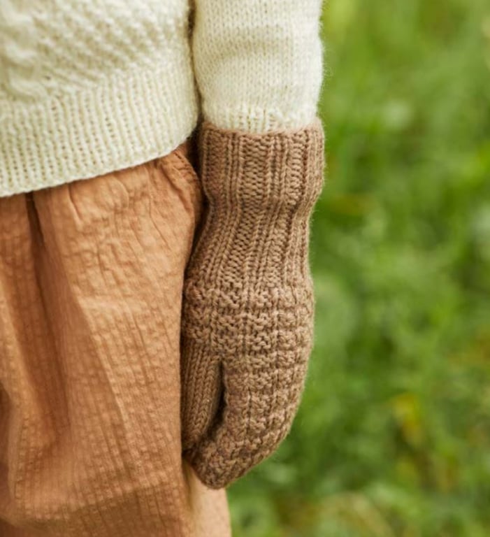 En person iført lysebrun strikkede vanter og beige strikket sweater med brune bukser, stående foran en grøn baggrund.