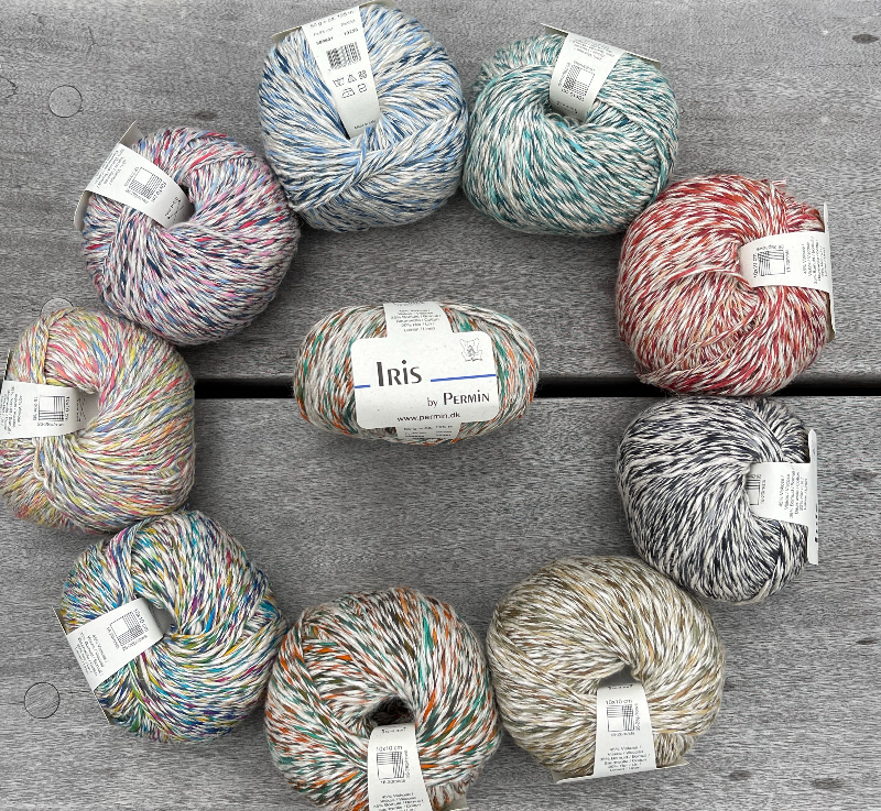 Ni klumper flerfarvet garn arrangeret i en cirkel på et gråt træbord, med en klump i midten mærket "Iris by Permin". (18)