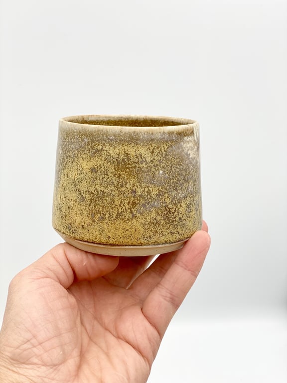 Hånd, der holder en lille, brunlig keramikkop med grovtekstureret overflade mod en hvid baggrund.